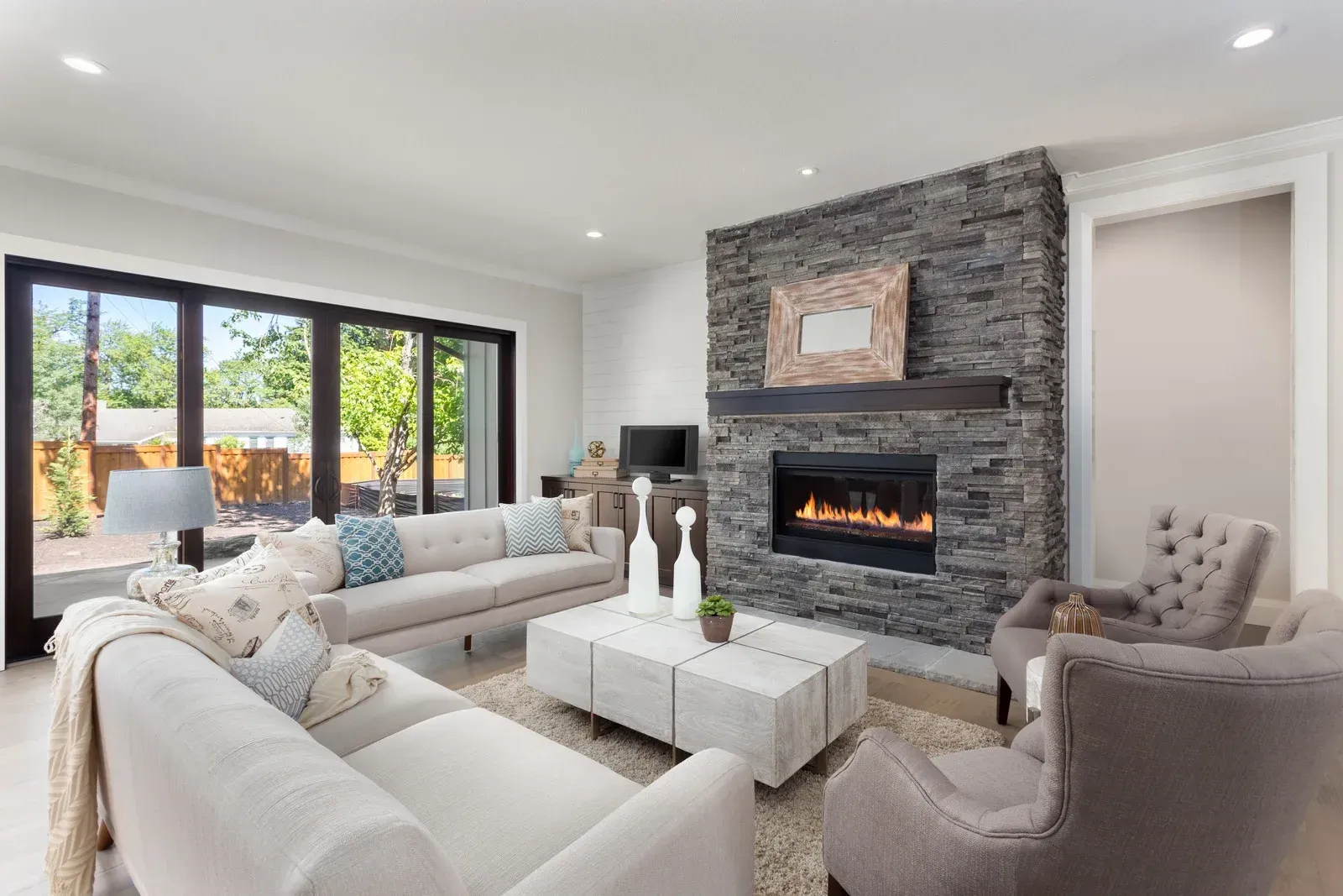 Bright modern living room with white sofas, stone fireplace, and large windows overlooking a patio