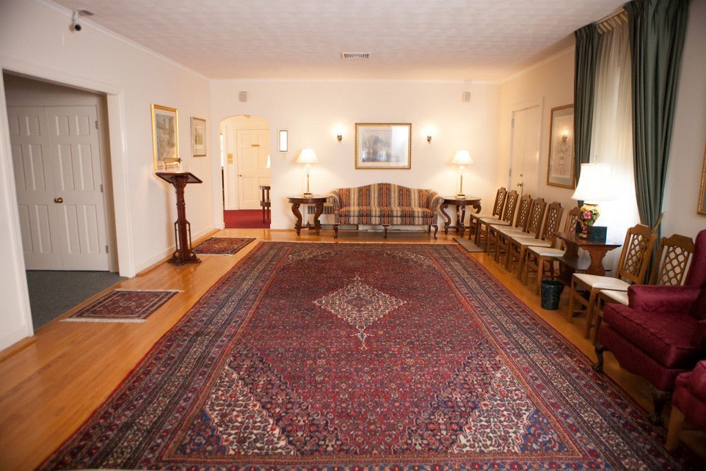 A room with a large red rug, sofa, chairs, and lamps. Doorway and door on the left.
