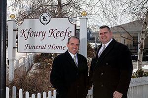Two men in suits stand in front of the Kfoury Keefe Funeral Home sign.