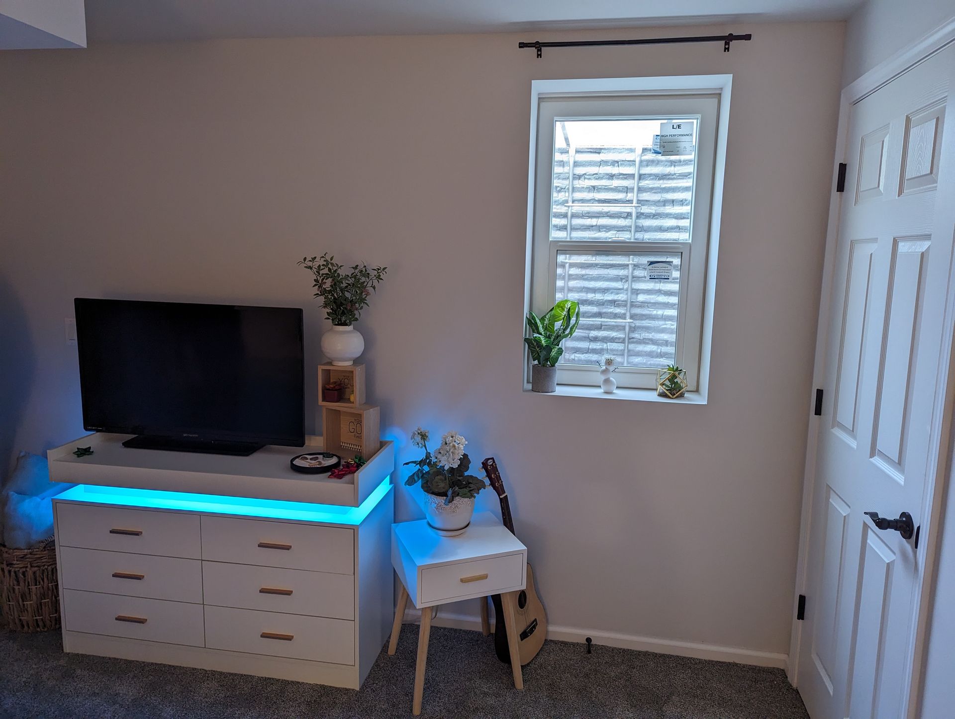 Bedroom with dresser, TV, window, and side table. Lighted dresser with a blue glow, white walls, and a door.