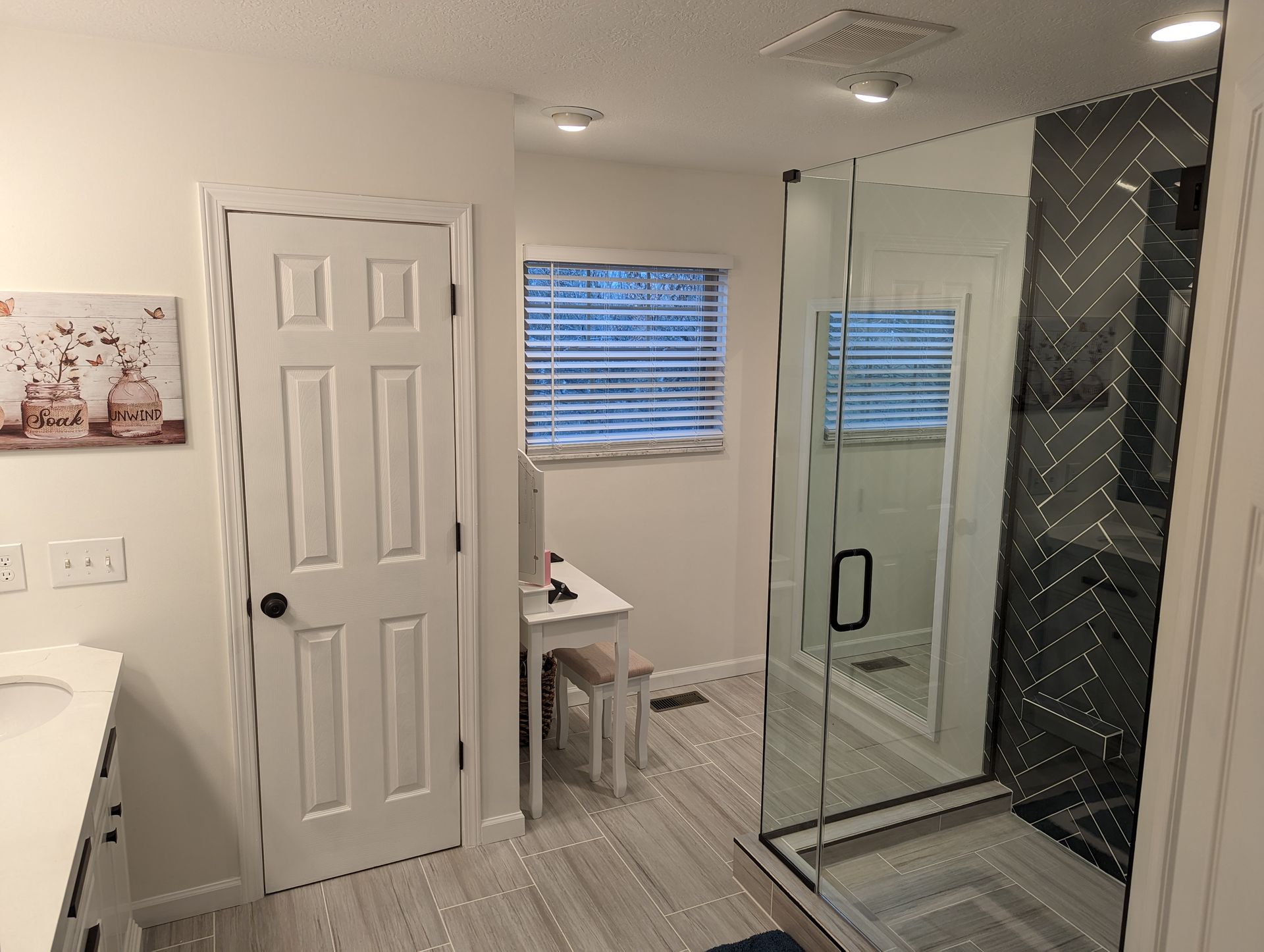 Bathroom with a white door, shower with black accents, and a window with blinds.