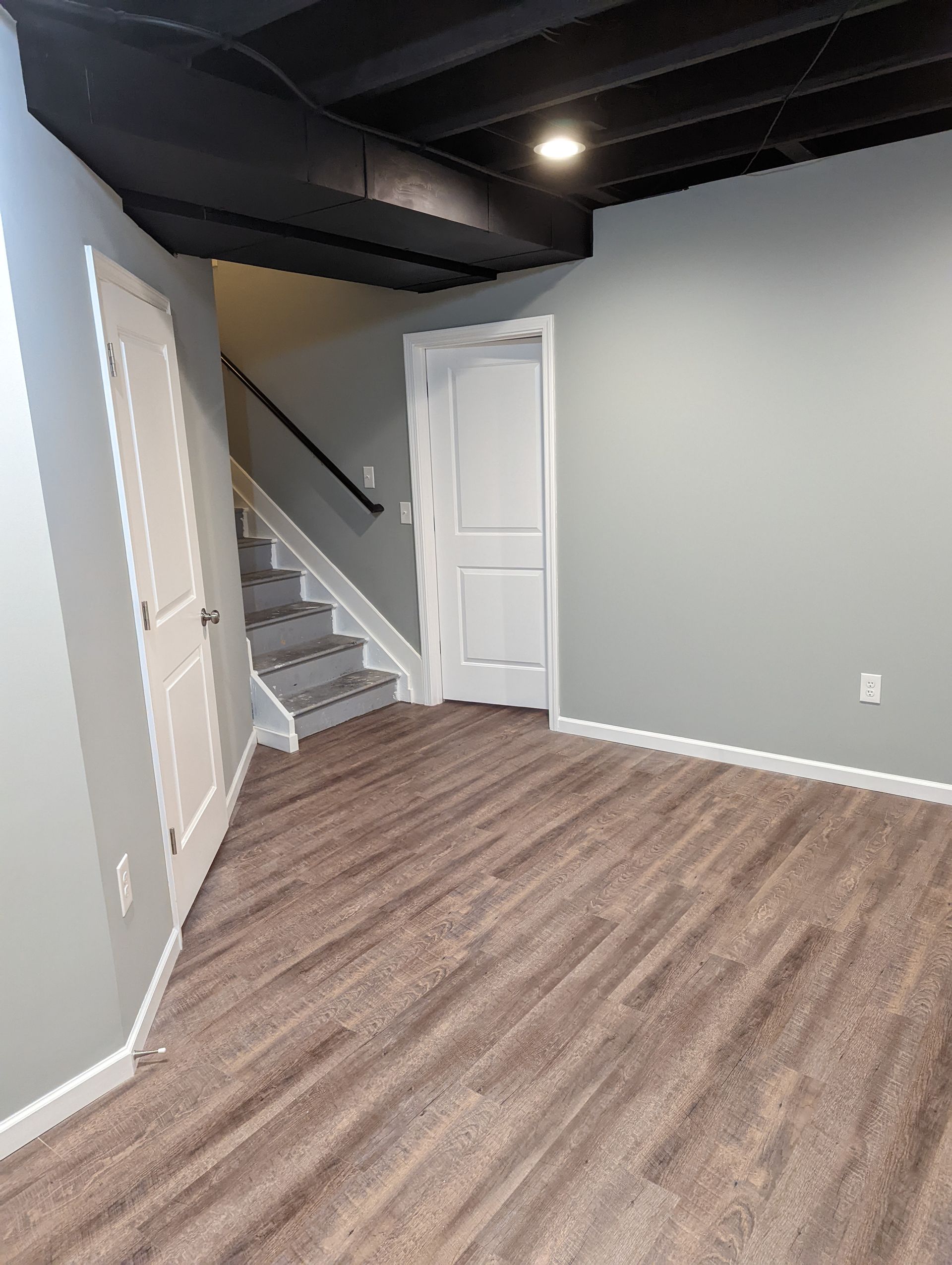 Finished basement with stairs, closed white doors, gray walls, brown flooring, and black ceiling.