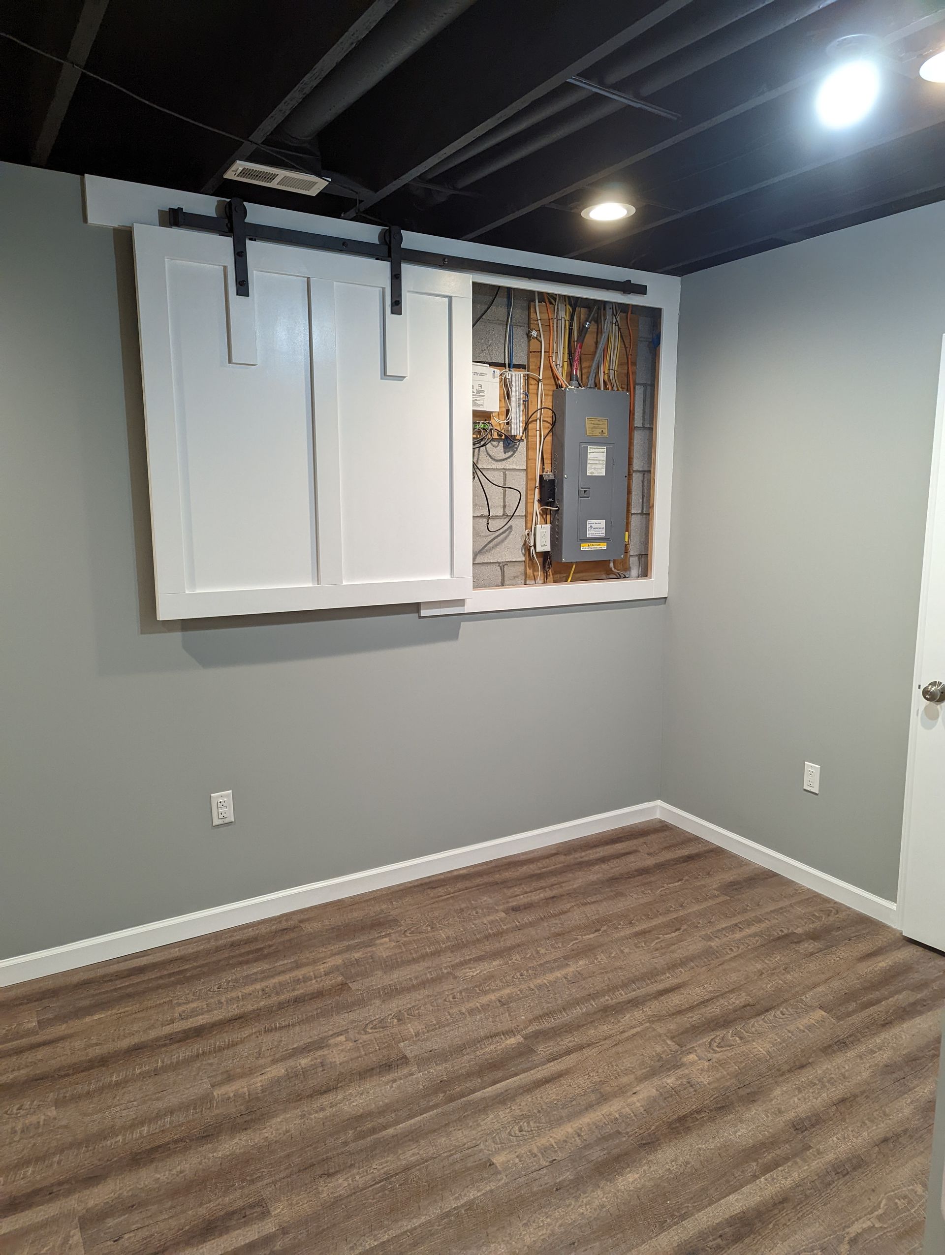 A room with gray walls, wood-look floor. White sliding doors cover an electrical panel. Black track hardware.