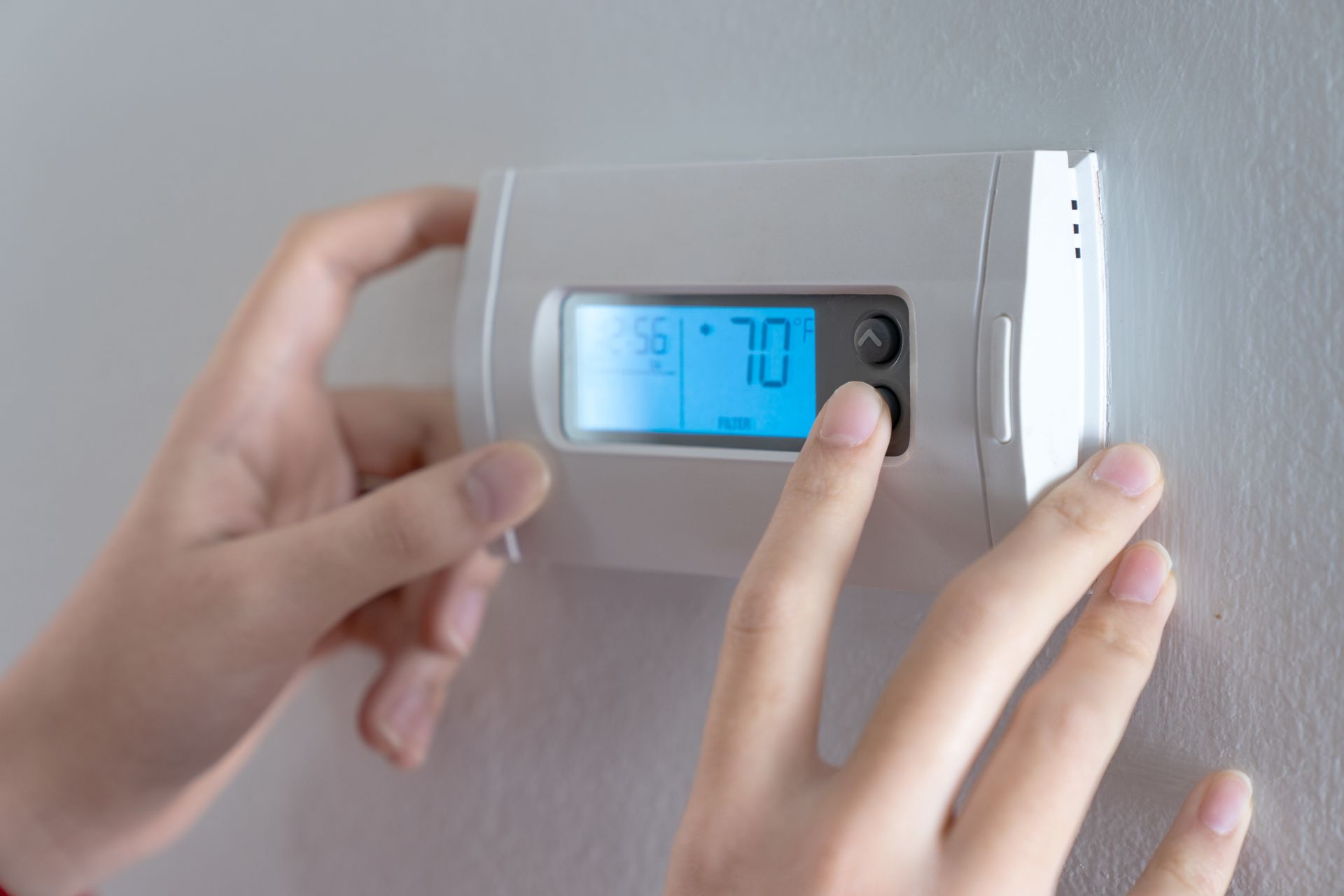 Hands adjusting a wall-mounted digital thermostat with a blue display. Hands adjusting a wall-mounted digital thermostat with a blue display.