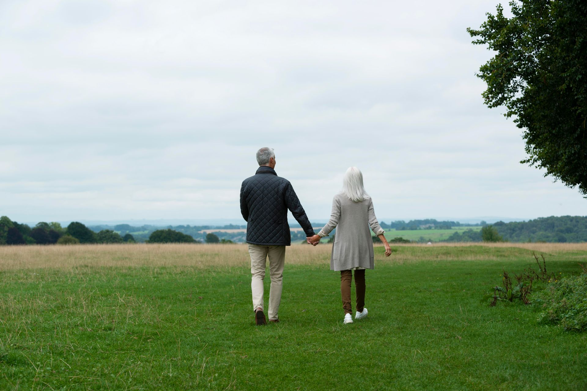 Couple walking hand-in-hand through a grassy field towards a distant treeline on a cloudy day.