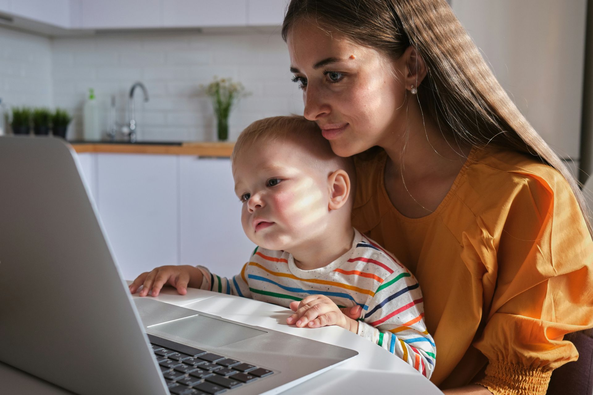 Woman and child looking at a laptop screen in a kitchen. The child is wearing a striped shirt, the woman an orange top.