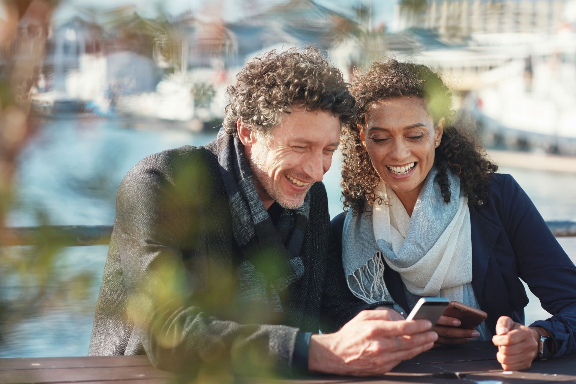 Couple smiles while looking at a phone outdoors at a waterfront, sunlit.