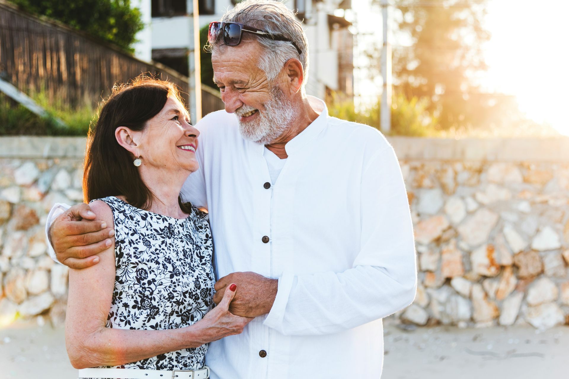 Smiling couple holding hands, man has arm around woman. They are outdoors near a stone wall, sunlit background.