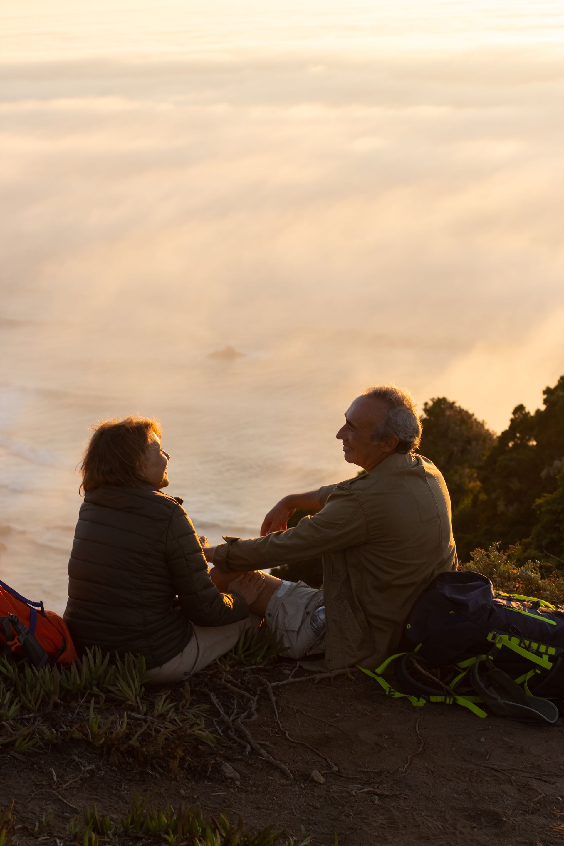 Couple sits on a hillside, holding hands, overlooking a sea of fog at sunset.