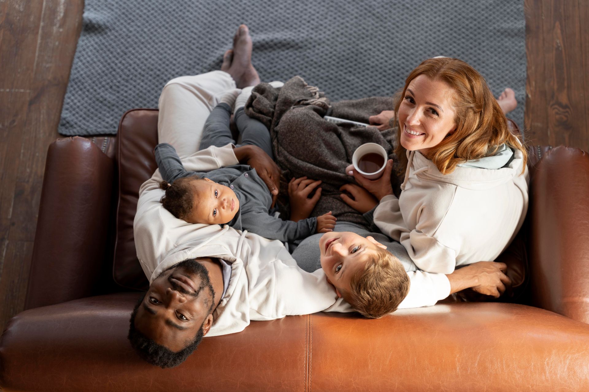 Family cuddles on a brown sofa, smiling. Gray blanket, cup of tea.