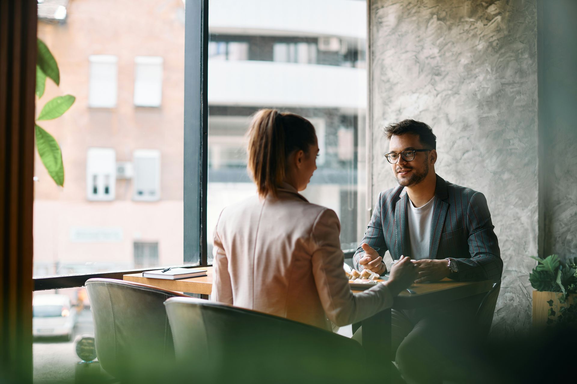 Two people seated at a table in a cafe, talking. Large window behind them.