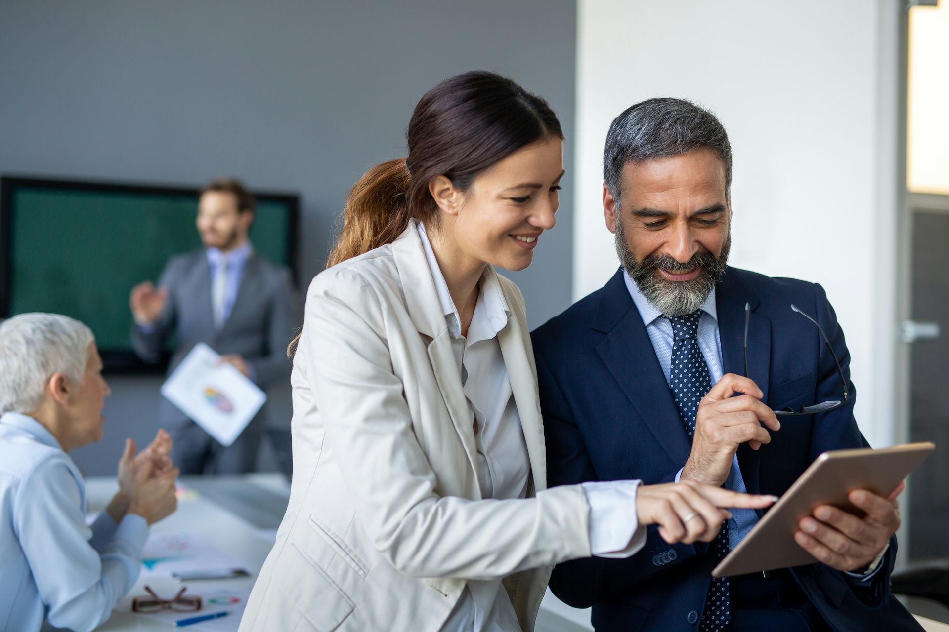 Two people looking at a tablet, smiling, in an office. Others in background.