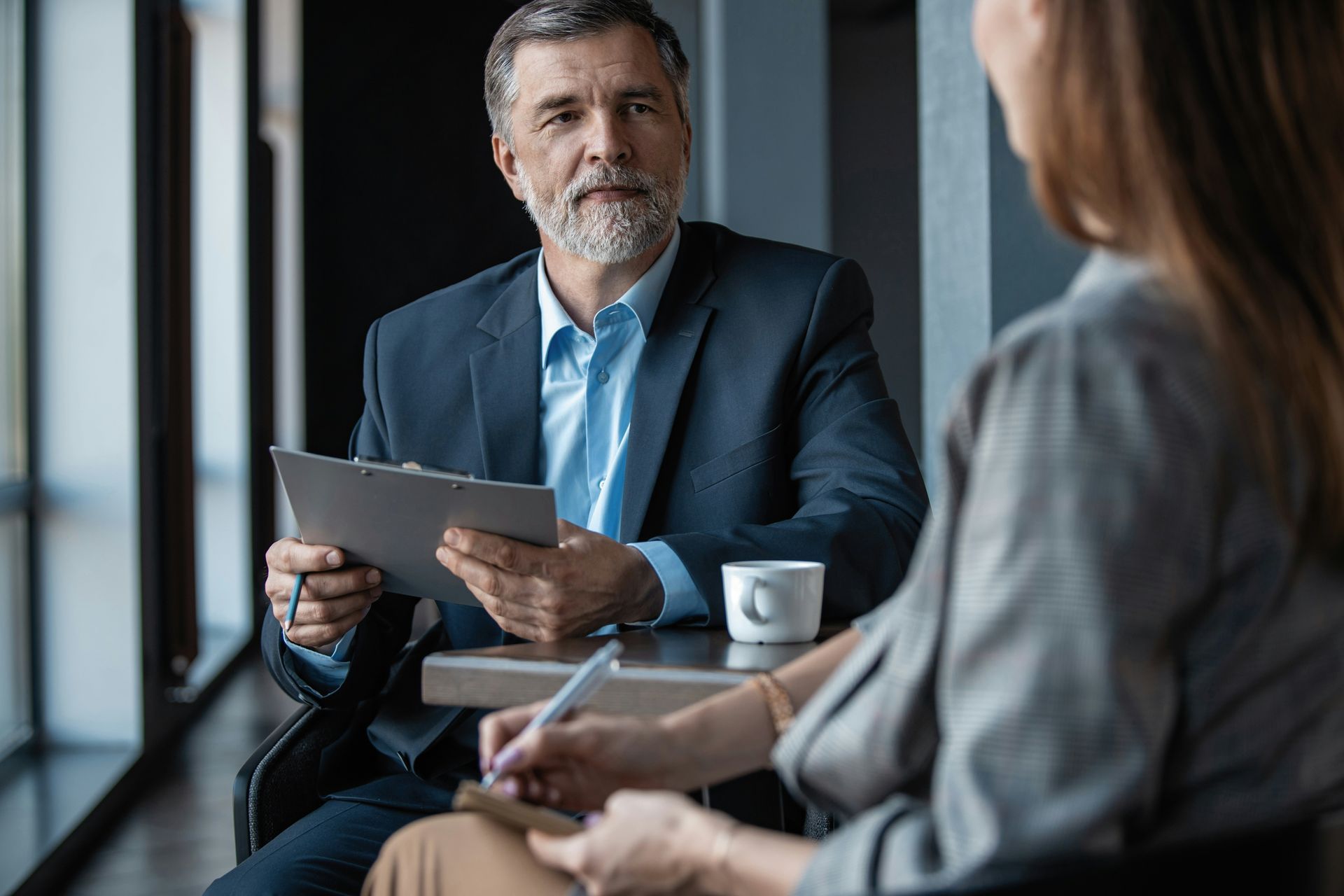 Man in suit, holding clipboard, and woman taking notes at a table. Coffee cup nearby.