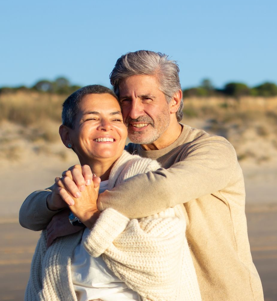 Couple embracing outdoors, smiling. Man hugging woman from behind; beach background.