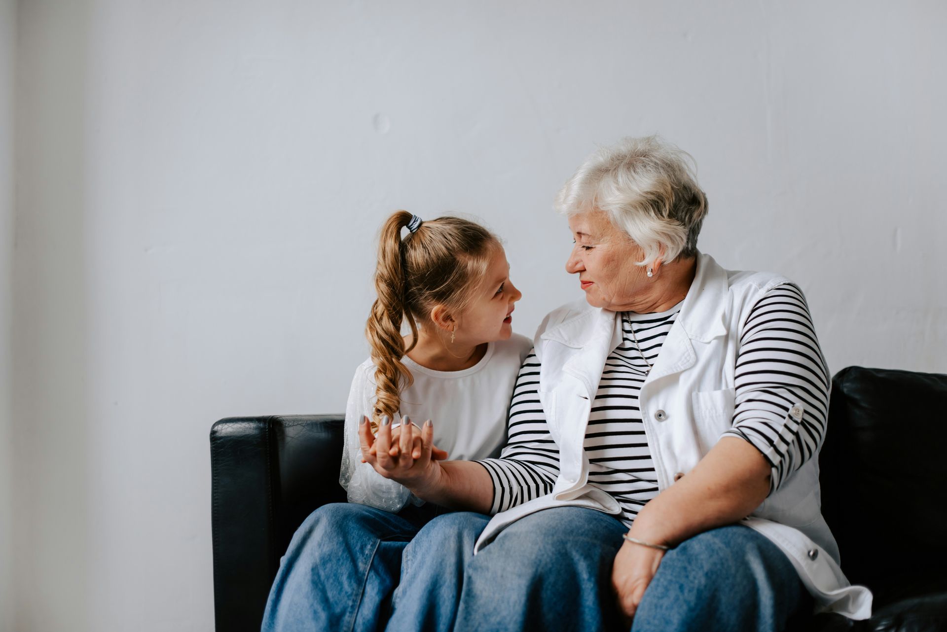 Grandmother and young girl sitting on a black couch, holding hands, and talking, against a white wall.