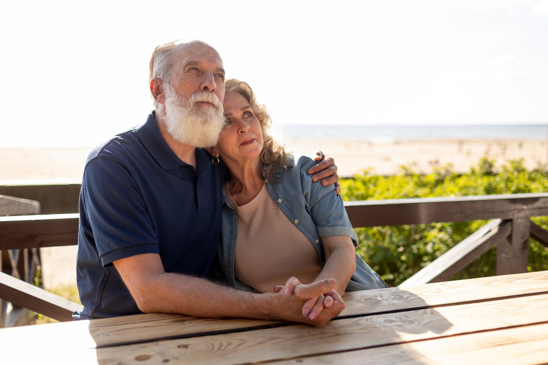 Older couple sits together at a table, looking up at the sky near a beach.