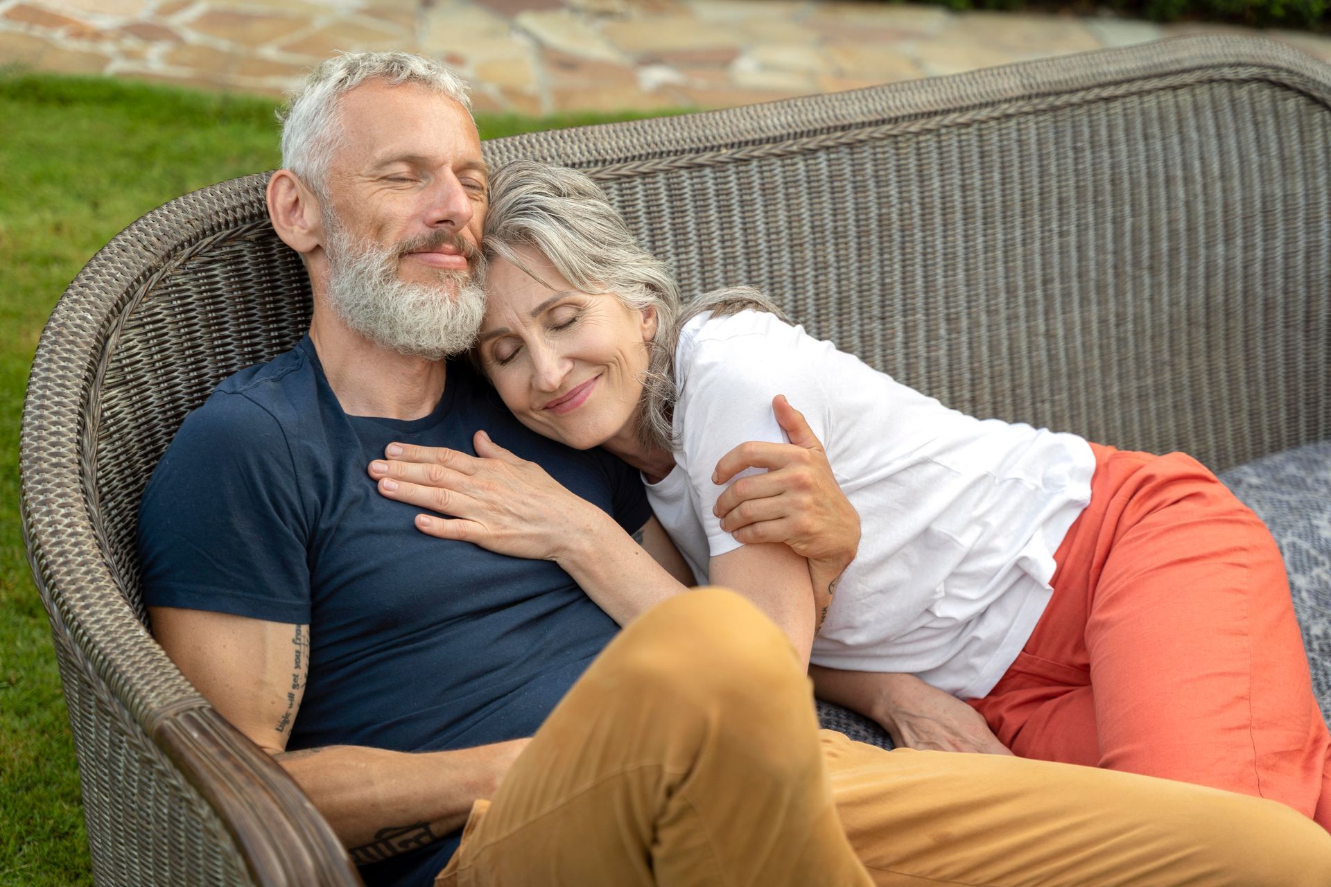 Couple embraces on wicker couch outdoors; woman rests head on man's chest; both have closed eyes.