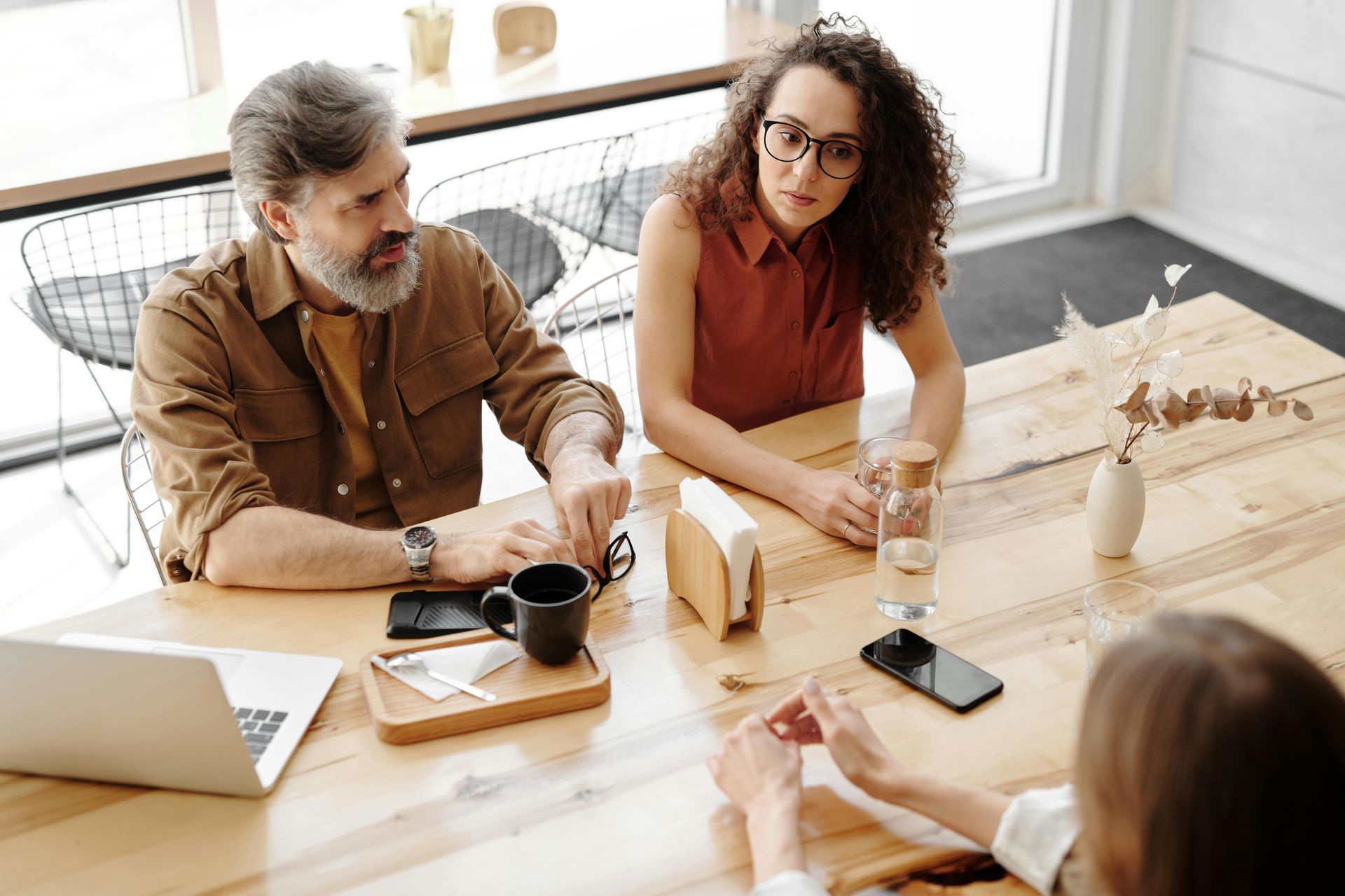 Three people seated at a wooden table in a cafe, engaged in a serious discussion, with a laptop and coffee.