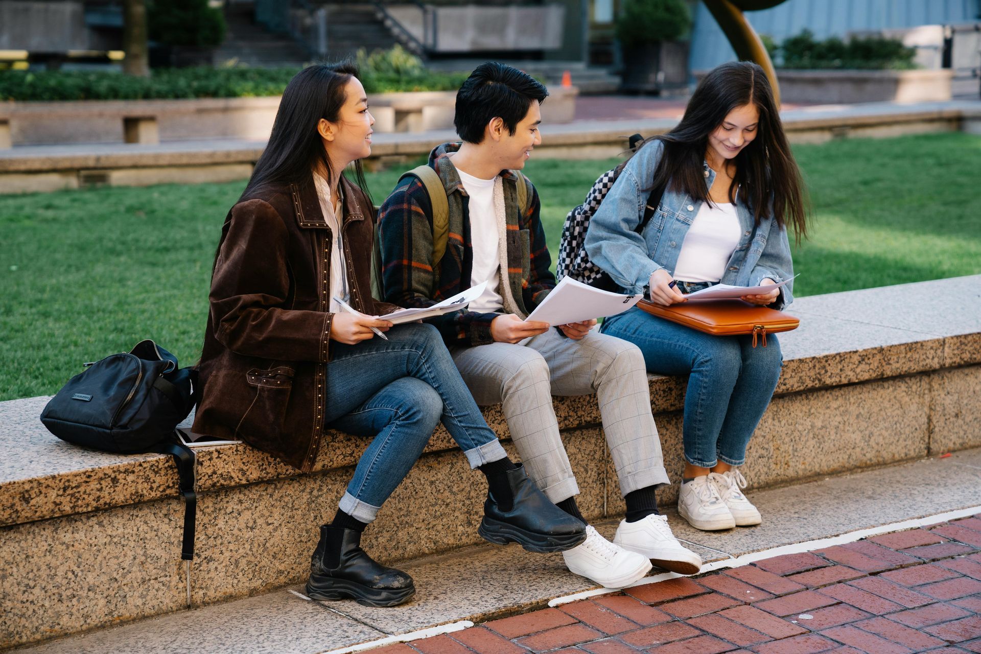 Three students sitting on a wall, looking at papers. Two women, one man, outdoors, sunny day.
