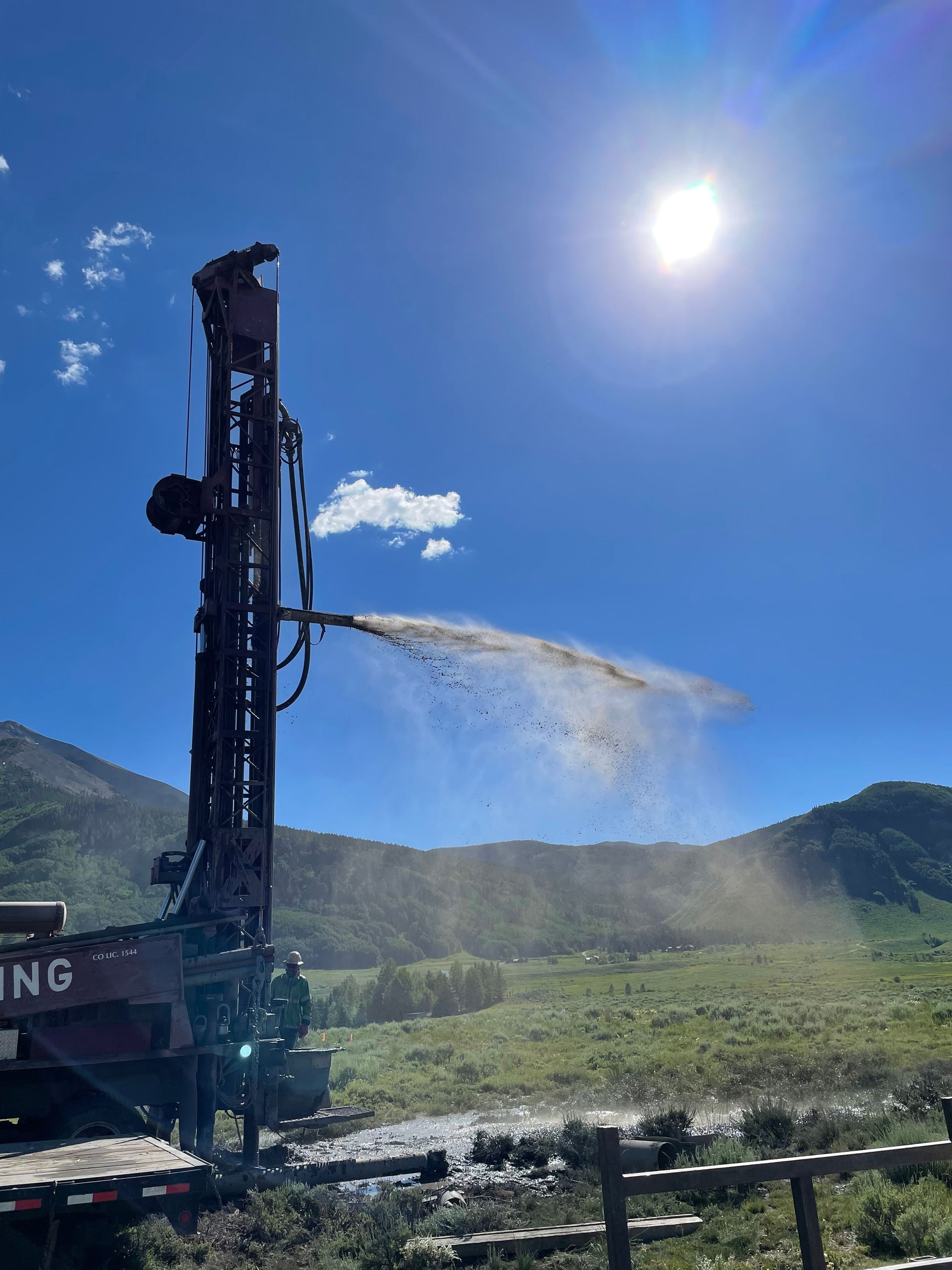 A water well is being drilled in a field with mountains in the background.