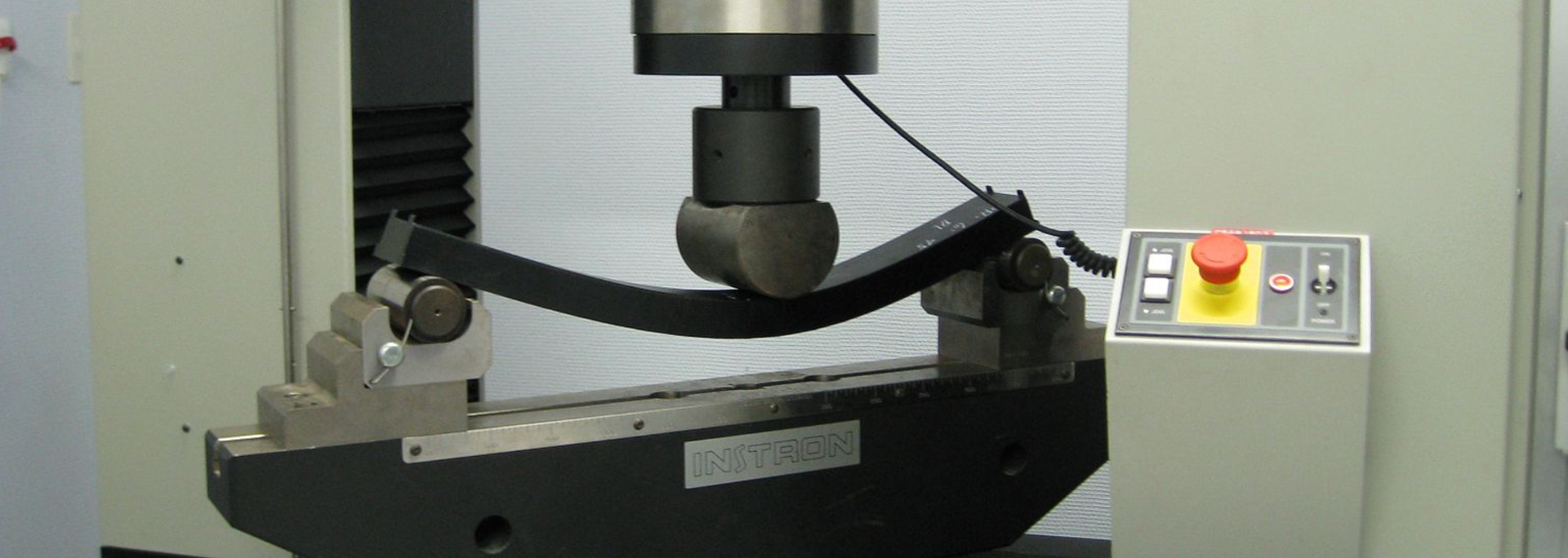 A machine testing the flexural strength of a black material sample. A red button is visible on the control panel.