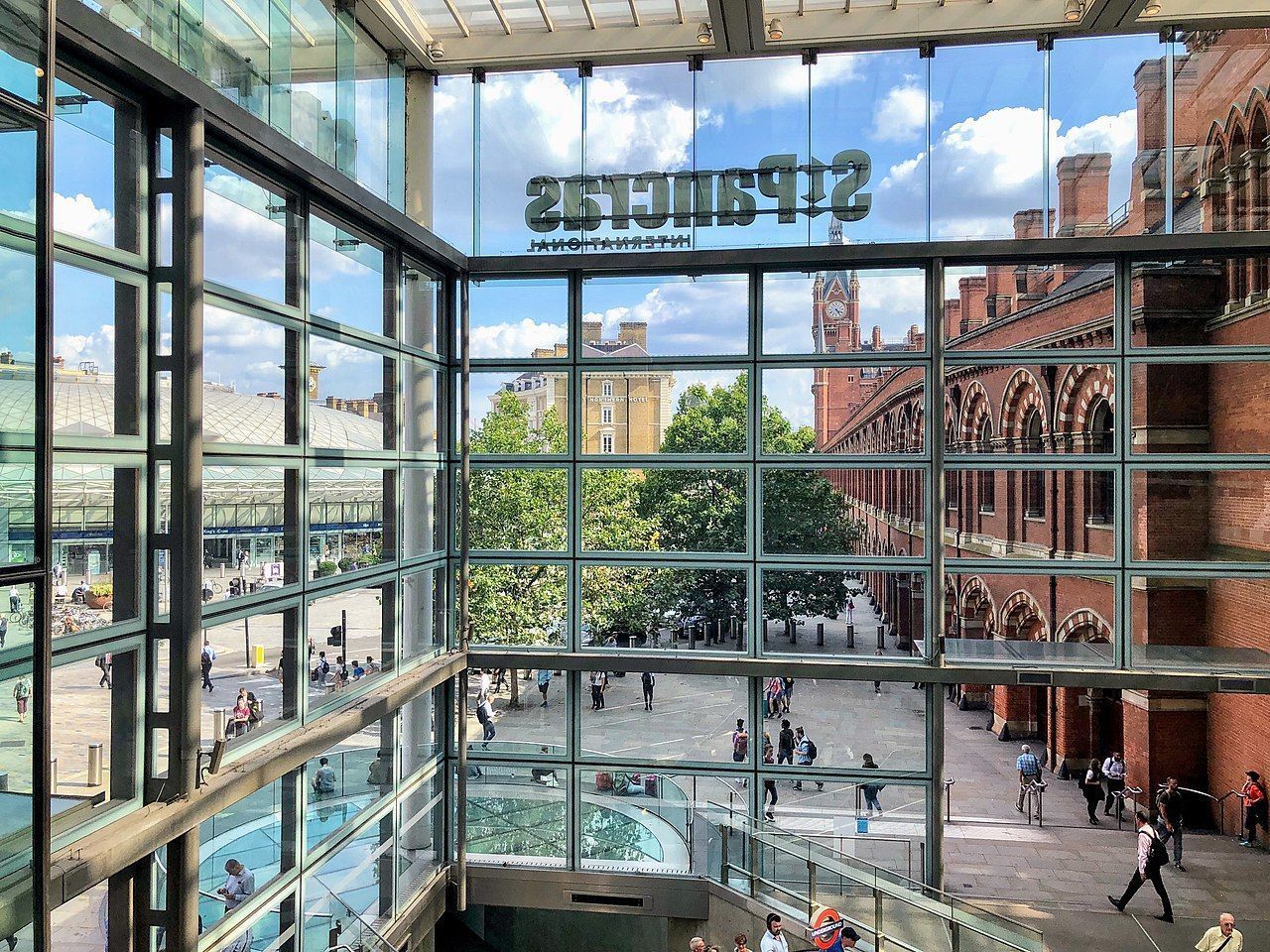 View of St. Pancras station exterior through glass windows. People walking, brick building with clock tower visible. Sunny day.
