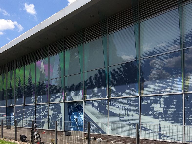 Large glass facade with mirrored images of a street and greenery, reflecting the blue sky.