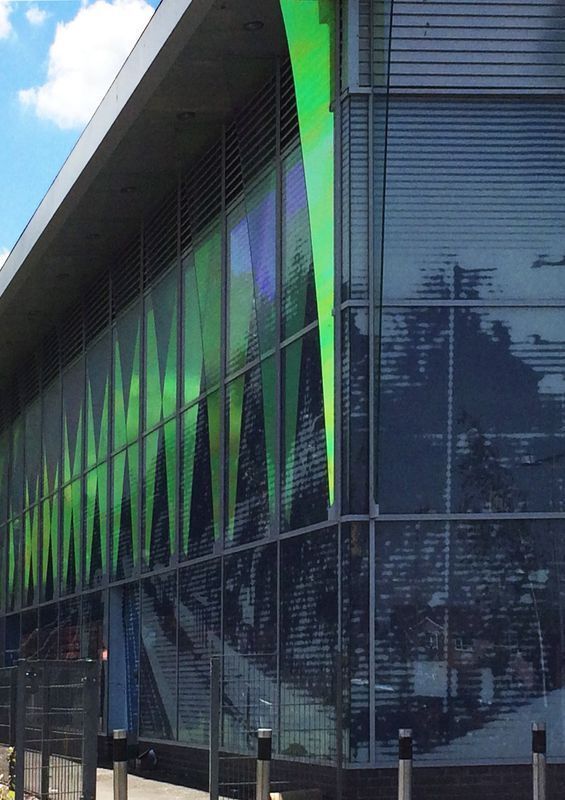 Modern building with green and blue glass panels, reflecting trees.