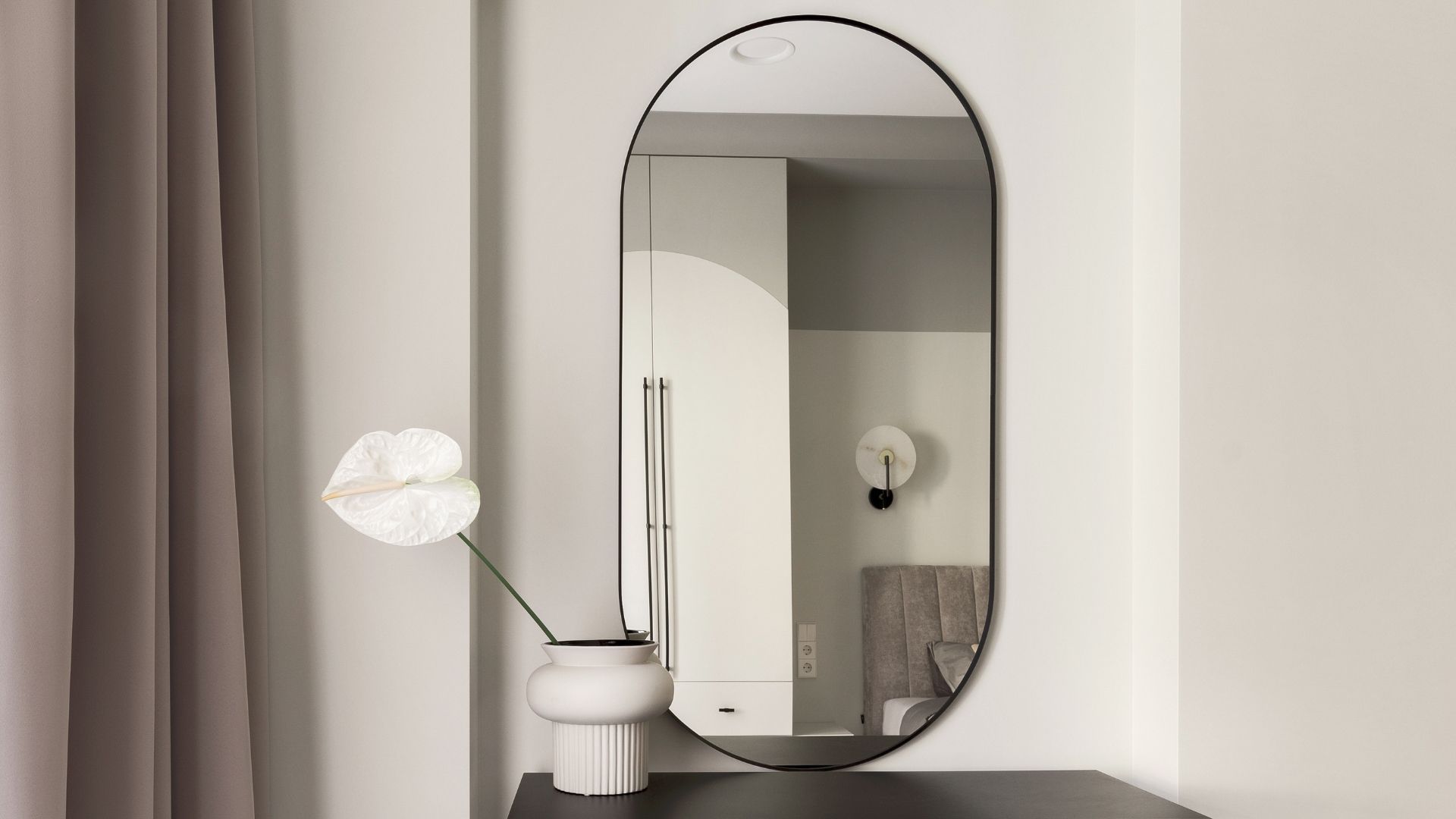 Oval mirror reflecting a cream-colored room with a flower vase on a dark surface.