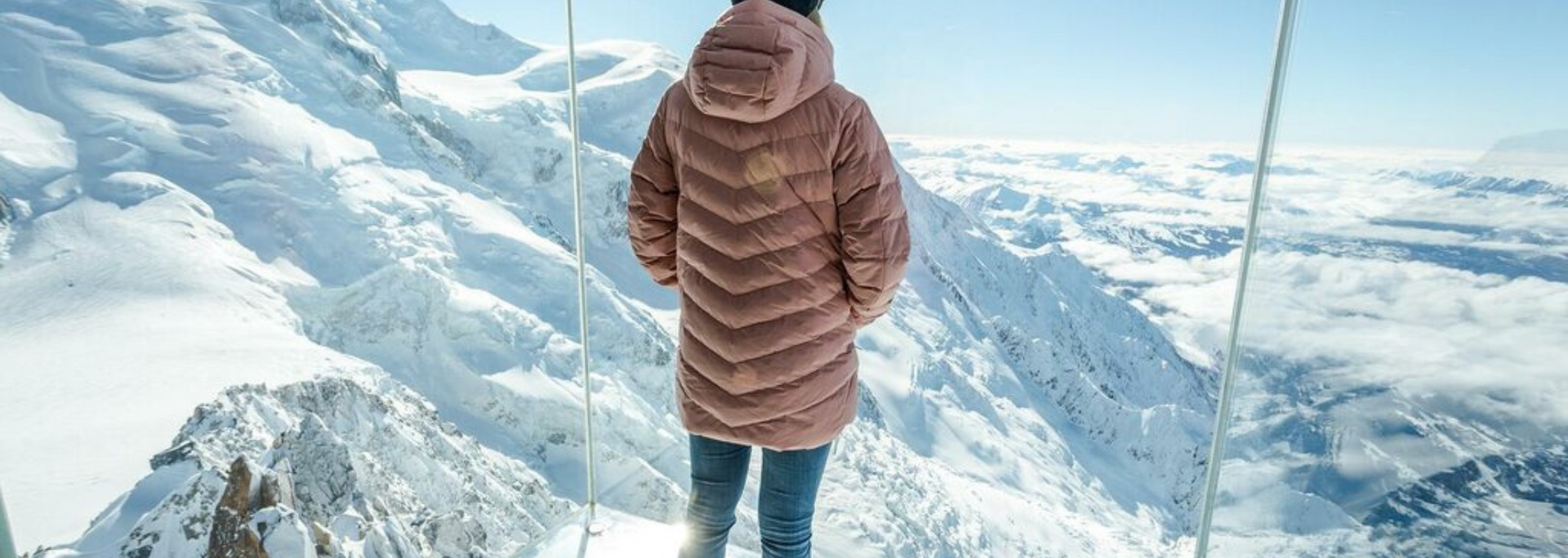 Person in pink coat standing before snow-covered mountains, view from a glass structure.