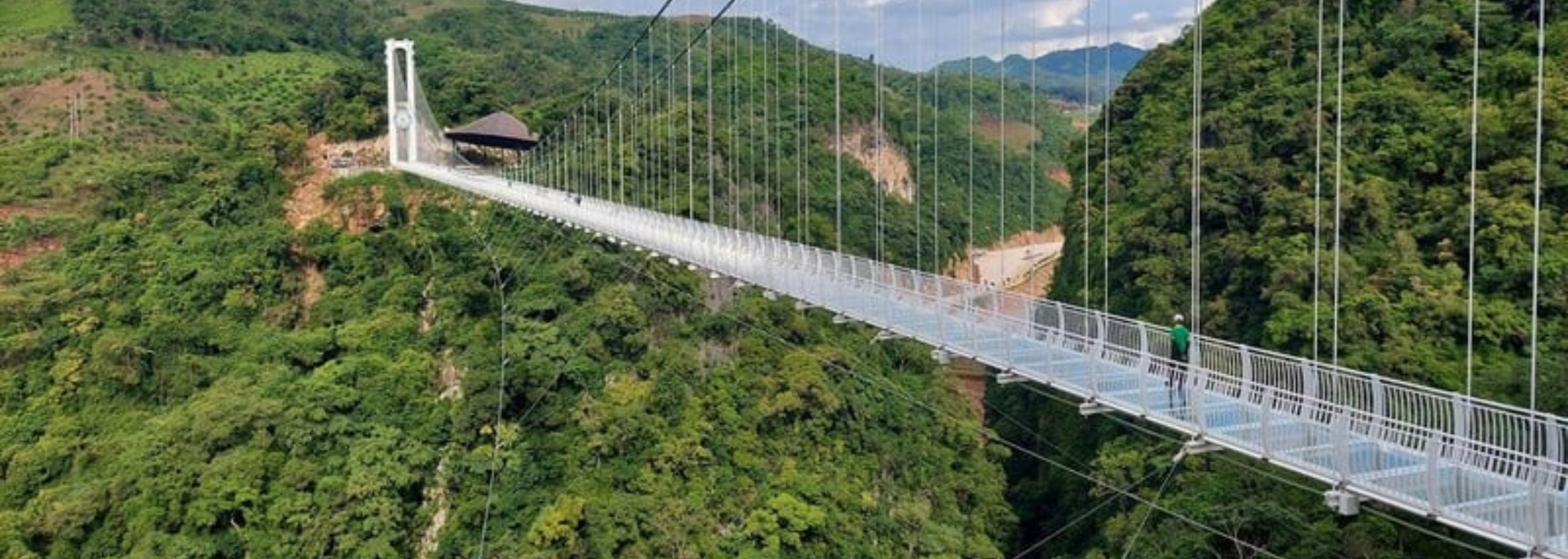 A long glass-bottomed bridge suspended high above a lush green valley. The bridge leads to a distant white tower.
