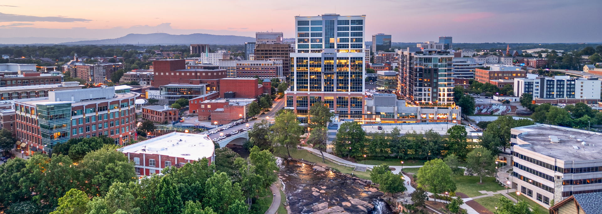 City skyline with tall buildings, trees, and mountains in the background at sunset.