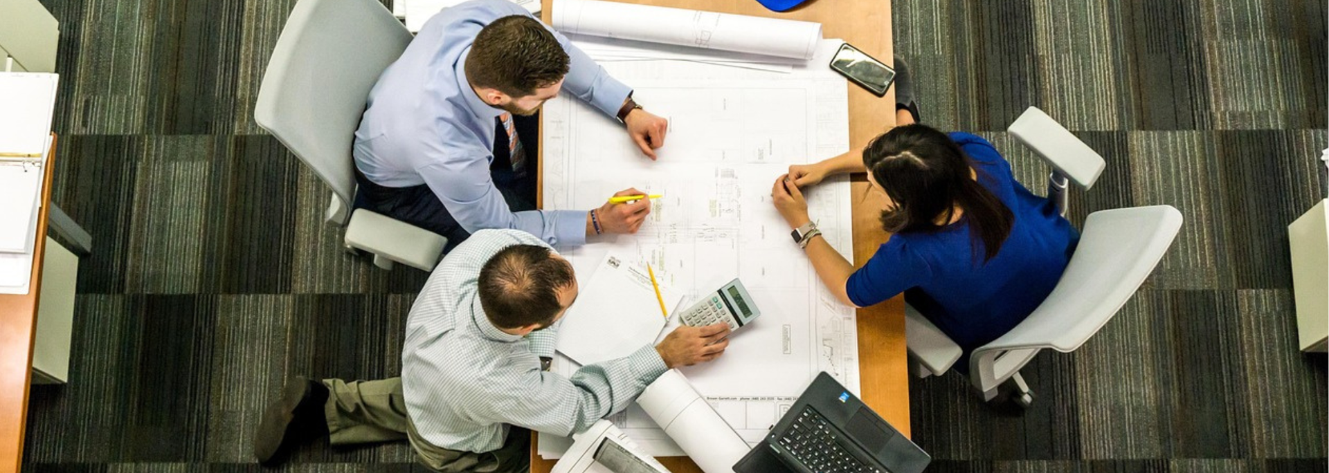 Team of people collaborating around a table, reviewing blueprints in an office setting.