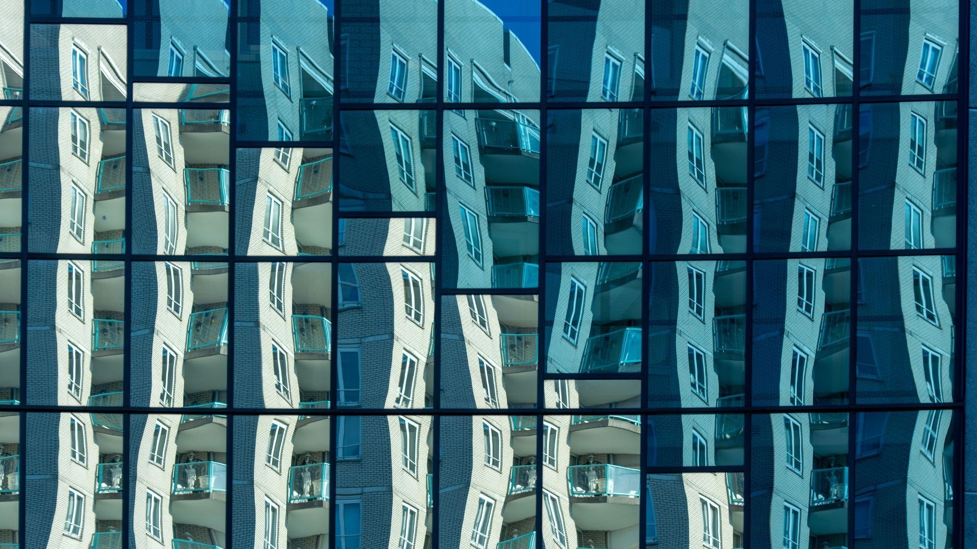 Glass building facade reflects another building with balconies, blue hues.