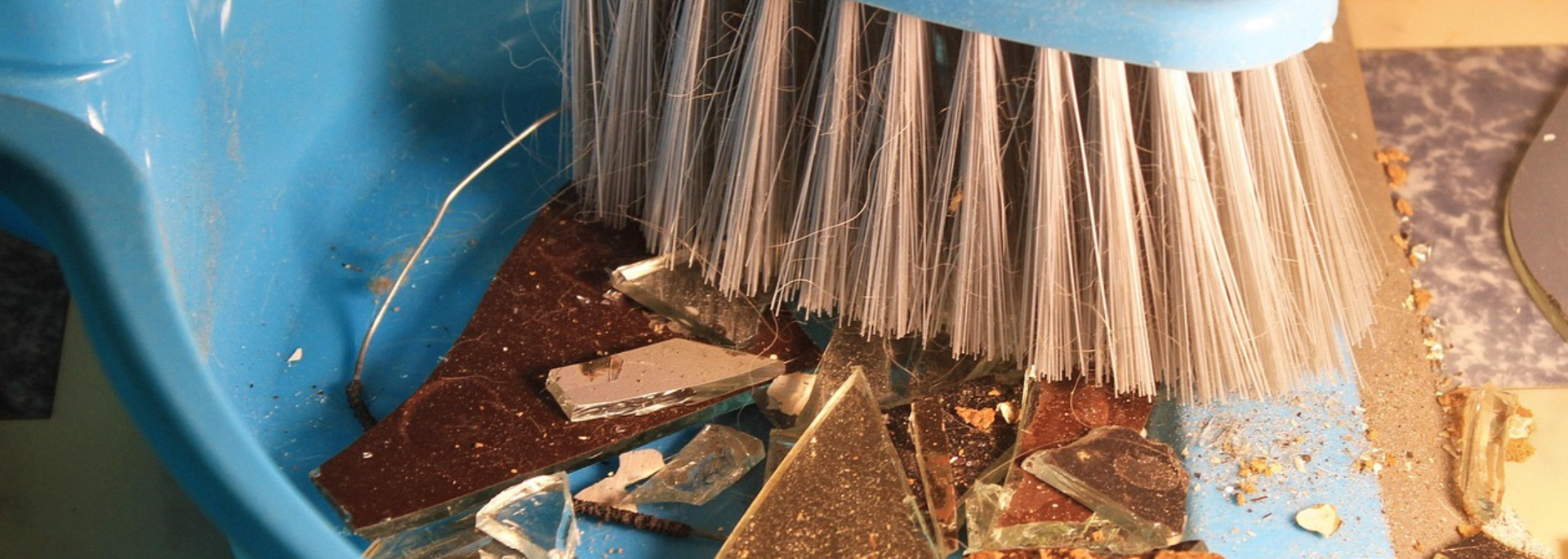 A blue dustpan collects shattered glass pieces as a brush sweeps them.