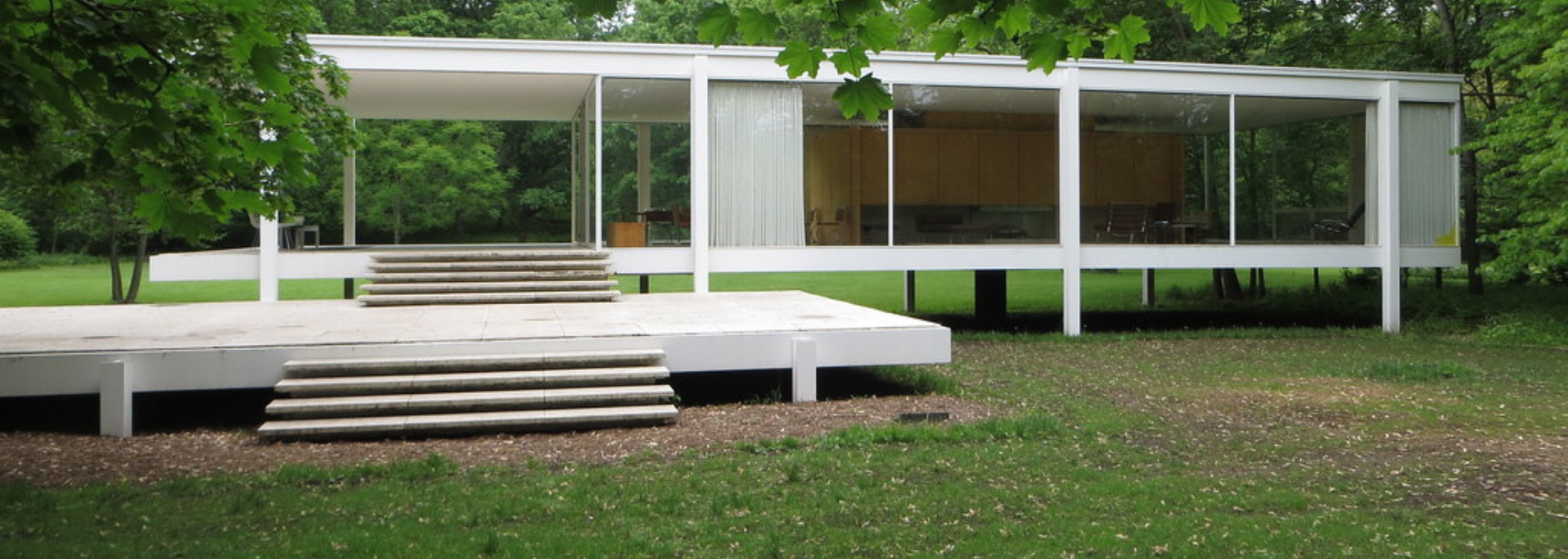 A white modern house with glass walls and a flat roof, set in a grassy area with trees.