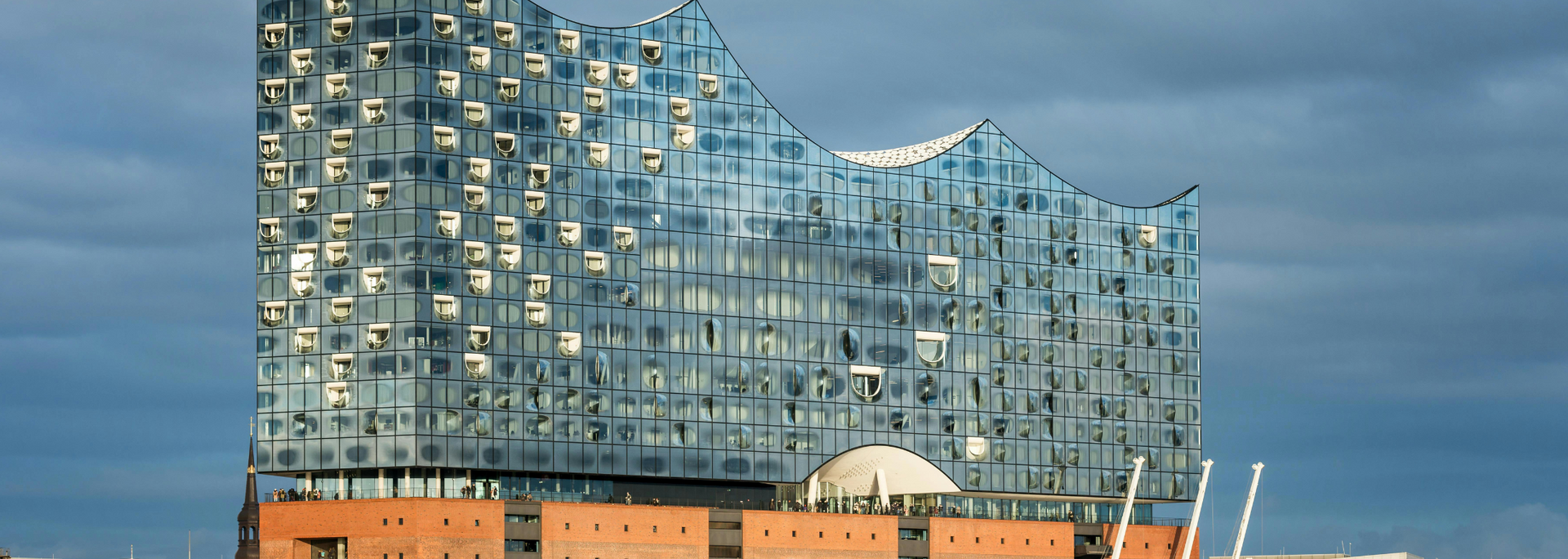 The Elbphilharmonie concert hall in Hamburg, Germany, with a glass facade and brick base, under a cloudy sky.