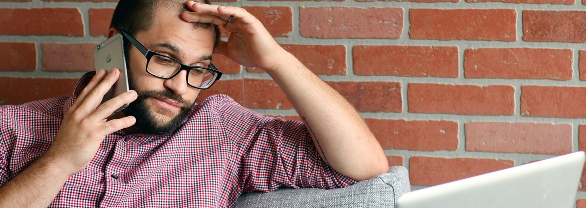 Man on phone looking stressed, leaning against brick wall, laptop visible.