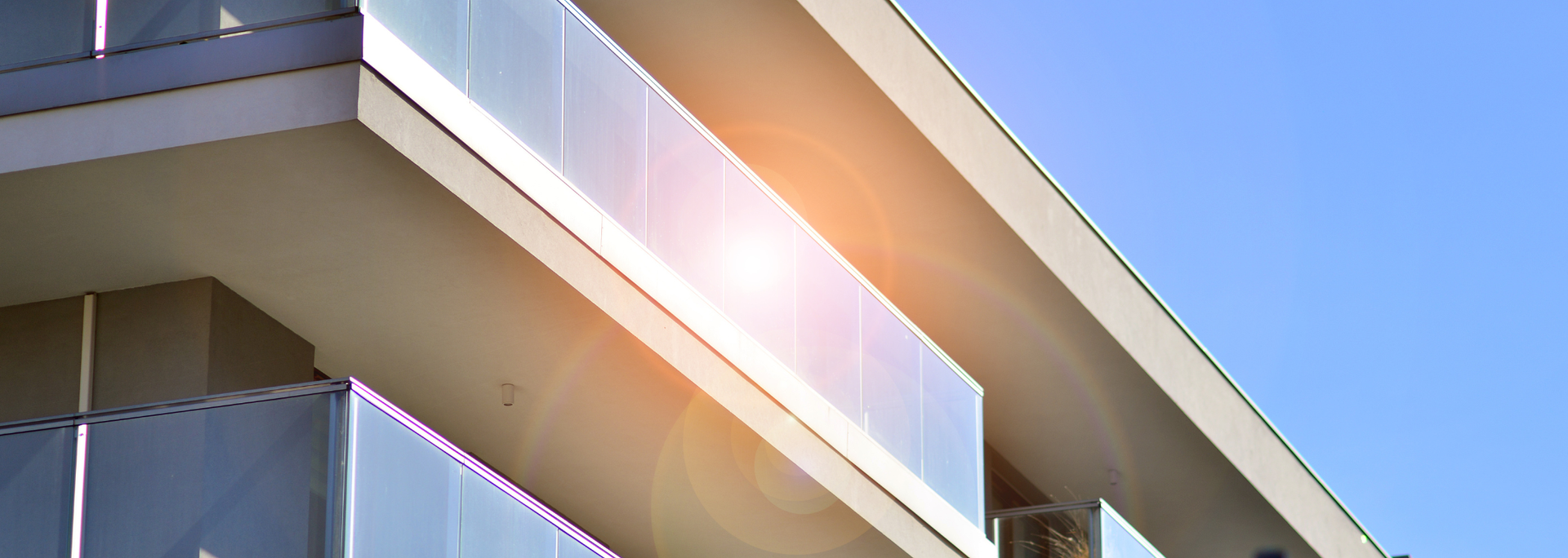 Modern building with glass balcony against a blue sky. Bright sun flare present.