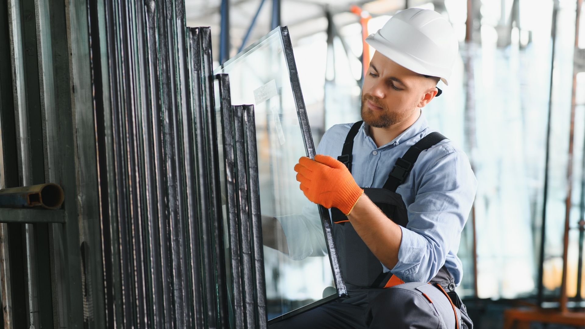 Man in hard hat and gloves inspecting glass sheets in a warehouse.