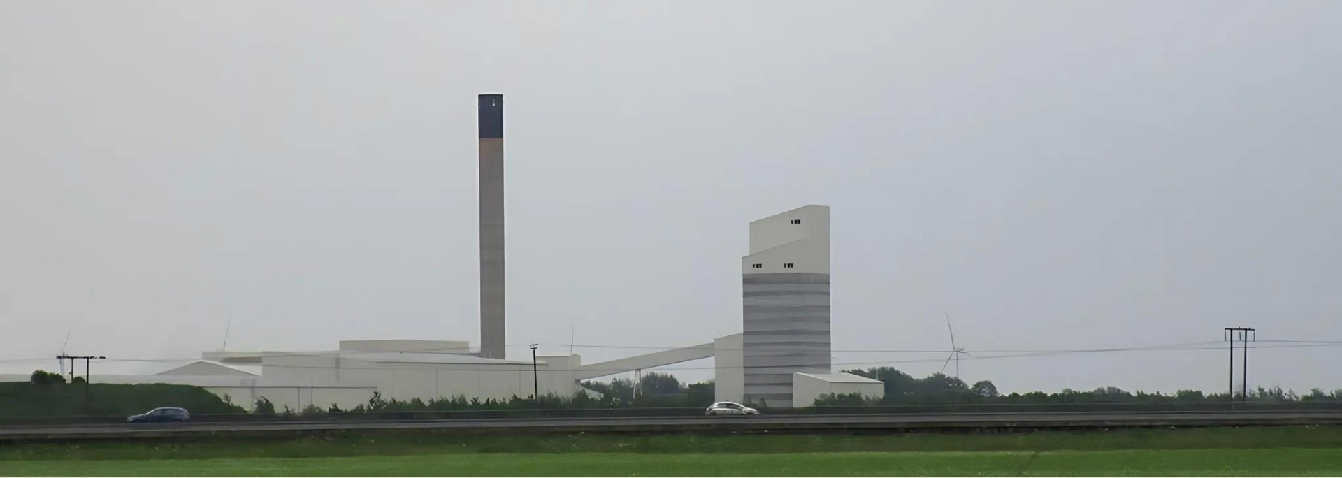 A large industrial complex with a tall chimney and a modern-looking building under an overcast sky.