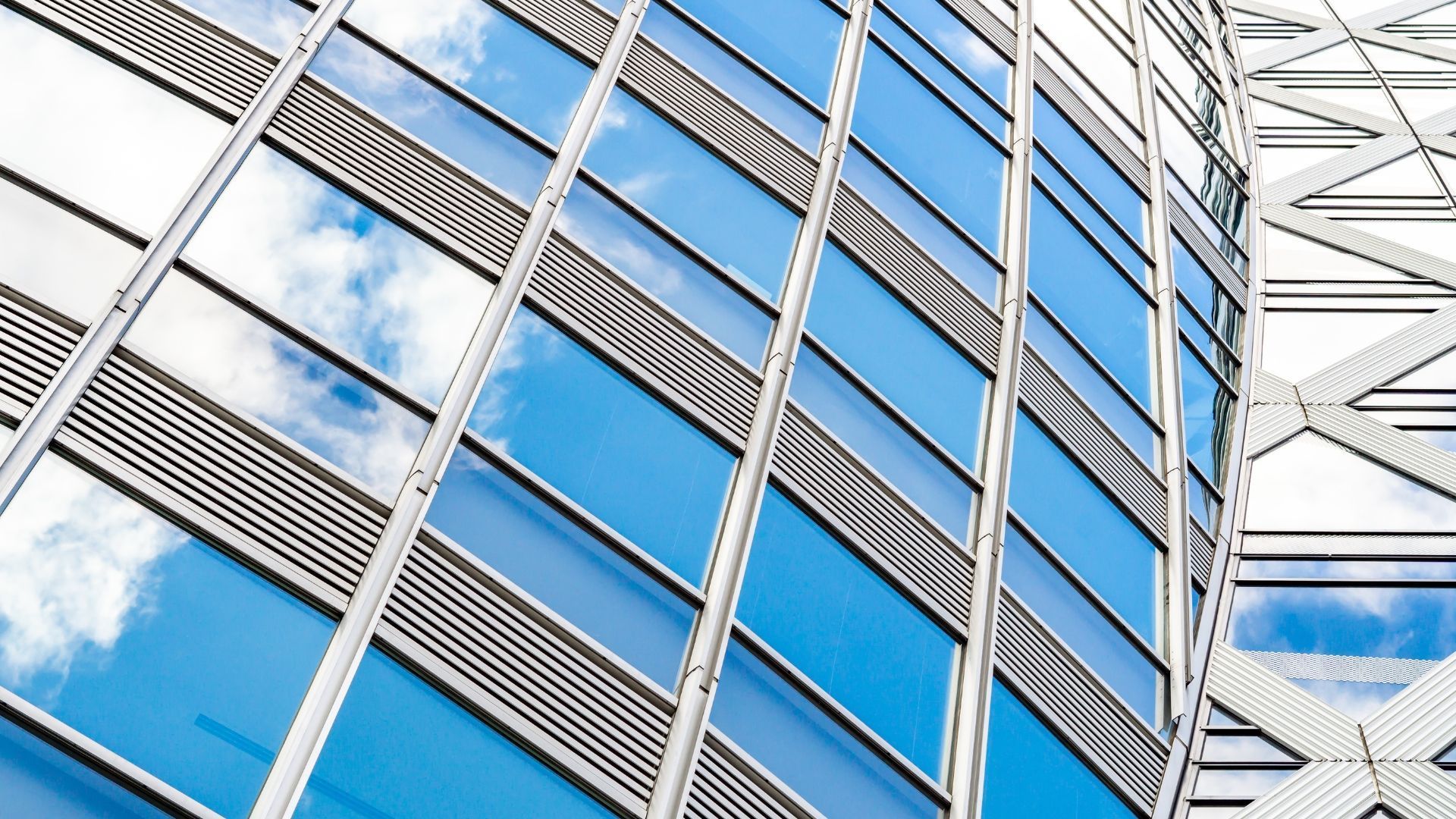Blue glass facade of a modern skyscraper reflecting sky and clouds, with a metal framework on the right.