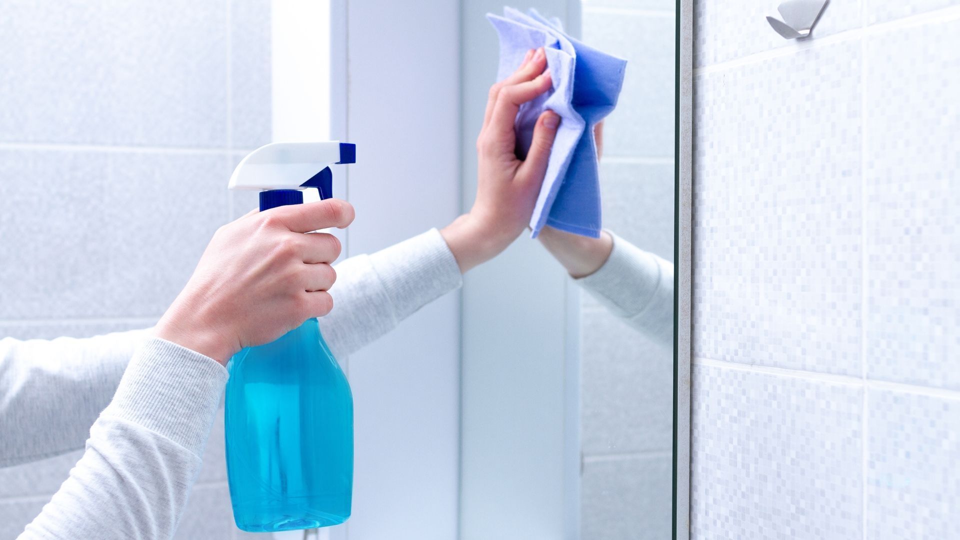 Person cleaning a glass shower door with spray bottle and blue cloth.