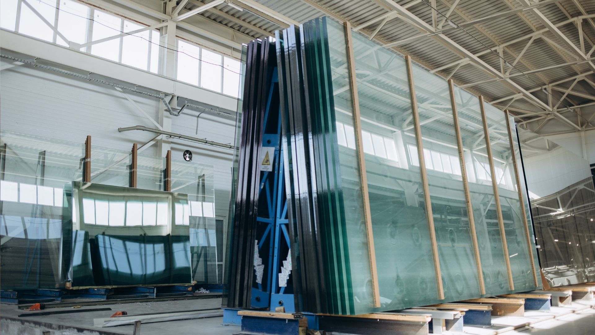 Large stacks of glass sheets in a warehouse setting, stacked on a blue cart for transport.