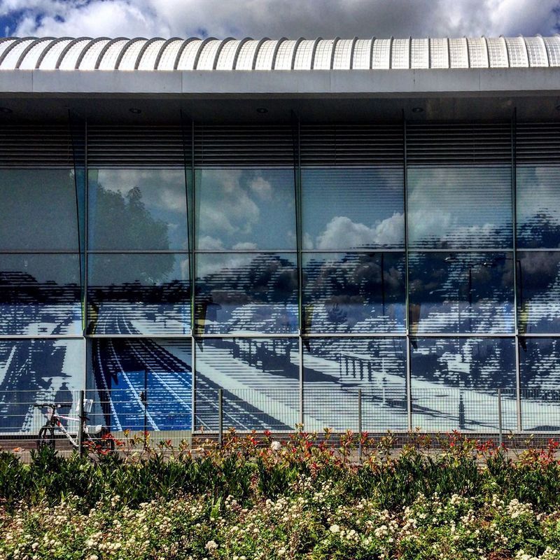 Exterior of a modern building with large glass windows reflecting a railway scene, cloudy sky above.
