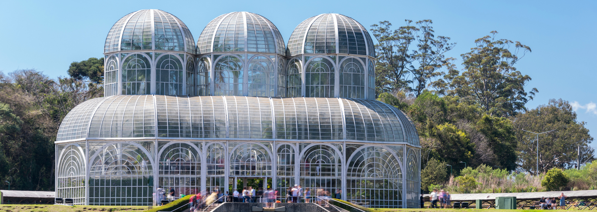 Glass greenhouse with domed roofs in a garden, under a bright blue sky.