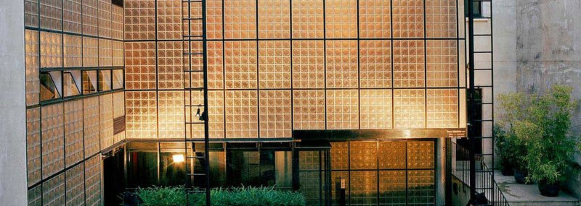 Exterior view of a building with glass block facade, metal frame, and greenery, illuminated with warm lighting.