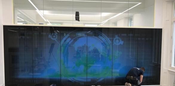 A person working on a large display screen showing a blue-green graphic, set inside a building.