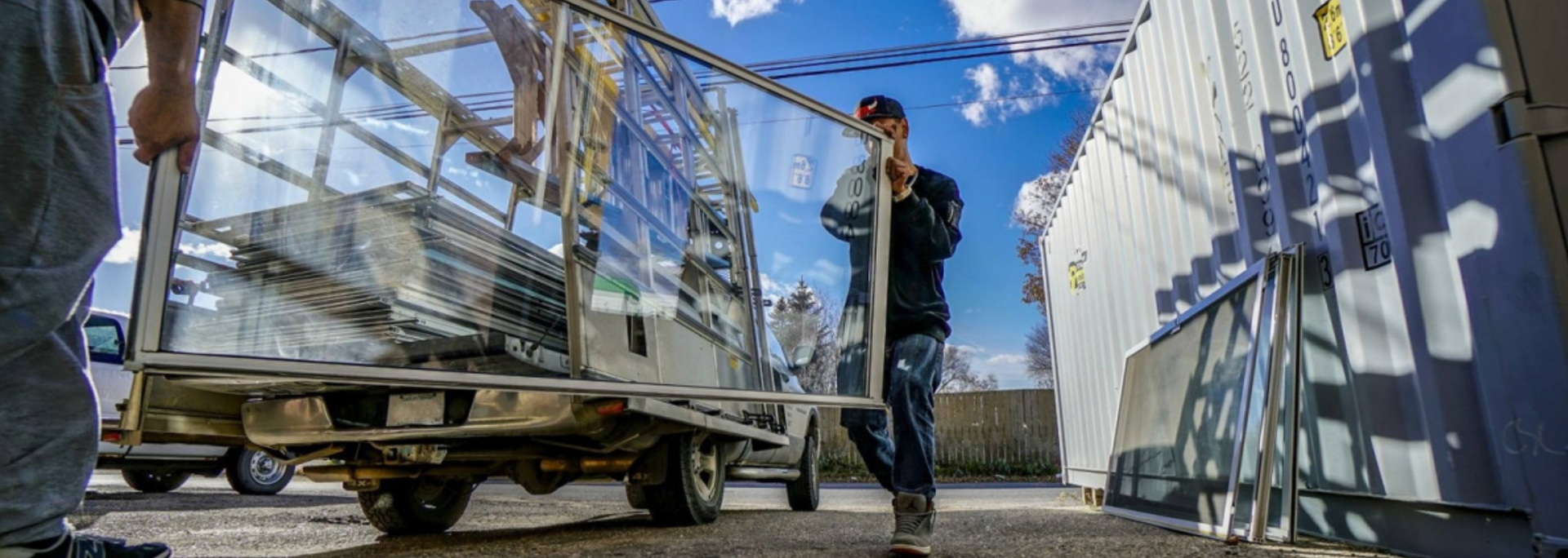 Two people moving large glass panes on a cart outside, under a blue sky.
