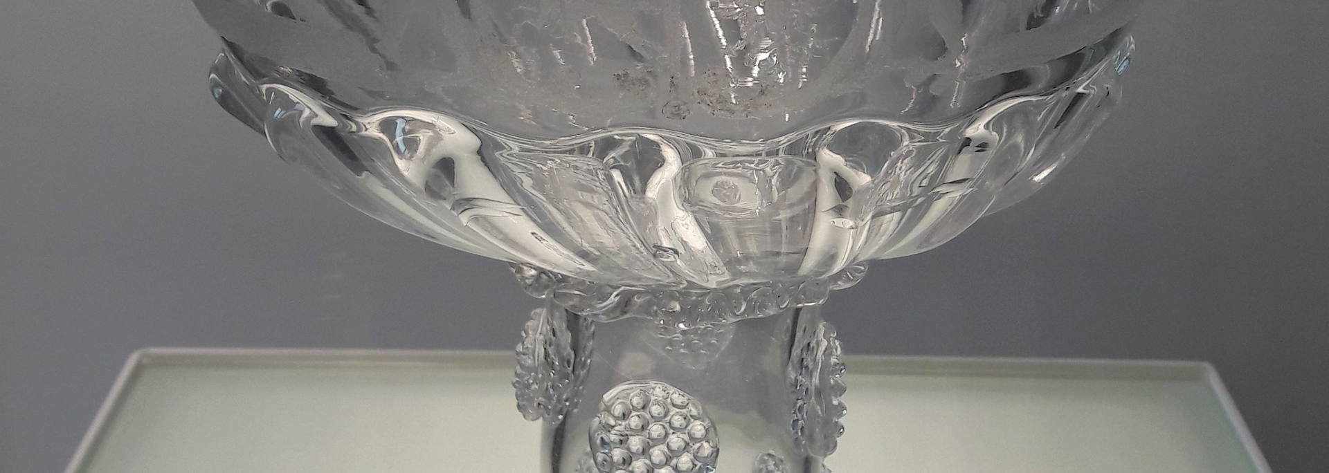 Close-up of a clear, ornate glass pedestal bowl. Details include textured stem and bowl rim with a floral design.